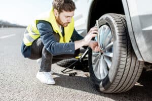 A man wearing a yellow safety vest is crouching next to a car on the roadside in Maricopa County, AZ, using a tool to check or fix the front tire—a clear example of roadside assistance in action.