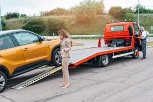 A woman in a suit stands near a yellow car being loaded onto a flatbed tow truck, receiving roadside assistance in Maricopa County, AZ, as the operator works at the back. The scene unfolds outdoors on a paved road.