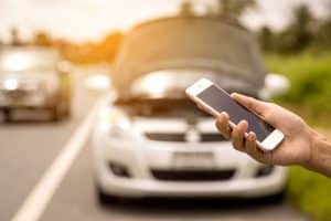 A person holds a smartphone near a white car with its hood open on the side of the road in AZ, likely calling for roadside assistance in Maricopa County due to a breakdown or emergency situation.