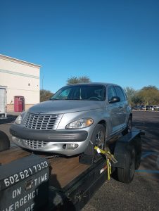 A silver Chrysler PT Cruiser is parked on a flatbed tow truck in a parking lot on a sunny day, showcasing efficient roadside assistance in Maricopa County, AZ. The background features a building, trees, and a clear blue sky.