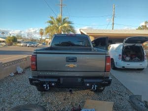A silver Chevrolet Silverado pickup truck, possibly used for roadside assistance Maricopa County, AZ, is parked in a driveway next to a white van with open back doors. Tools and boxes are scattered on the gravel, with palm trees and houses in the background.