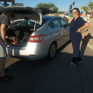 A woman in navy blue scrubs stands next to a silver car with the trunk open, smiling at the camera. Another person checks the trunk—perhaps they’re getting roadside assistance in Maricopa County, AZ, on a sunny day near a gas station.