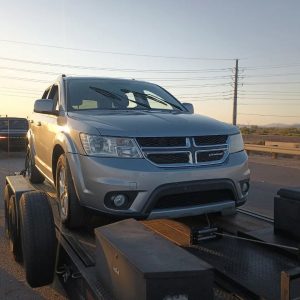A silver Dodge SUV, secured on a car trailer, awaits roadside assistance in Maricopa County, AZ. Parked on the roadside at sunset, power lines and a guardrail are visible in the background.