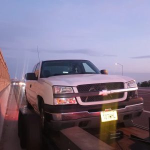 A white Chevrolet pickup truck is parked on a flatbed tow truck at dusk on the side of a highway, showcasing roadside assistance in Maricopa County, AZ. The yellow license plate reads "TOWHUL," with car headlights visible in the background.