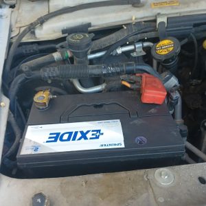 A close-up view of a car engine bay shows an Exide car battery with labeled terminals and surrounding engine components, including hoses and fluid caps—perfect for roadside assistance in Maricopa County, AZ.
