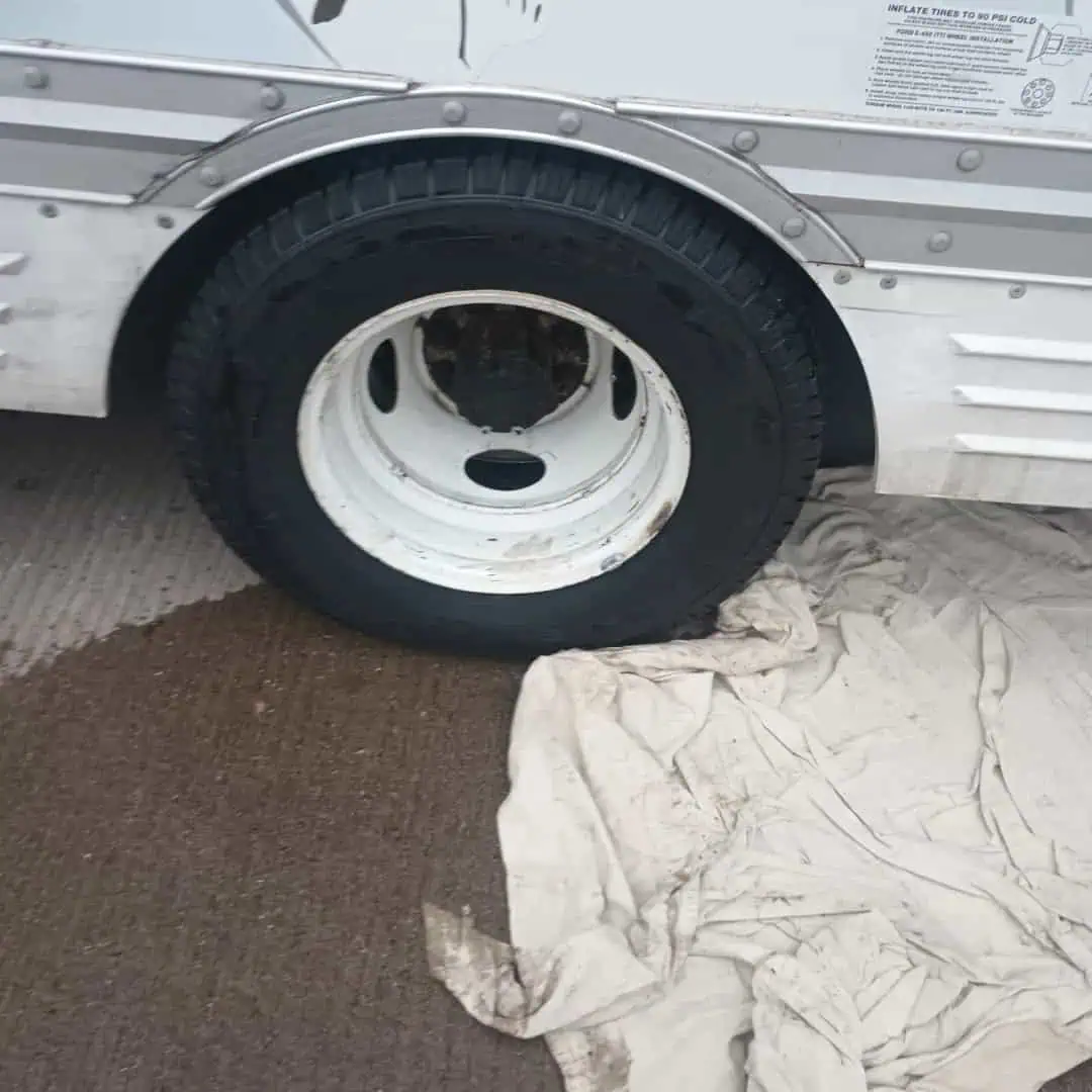 A large black tire on a white rim is attached to a vehicle, parked on a concrete surface. Part of the tire, possibly awaiting roadside assistance Maricopa County, AZ, rests on a dirty, crumpled white sheet spread on the ground.