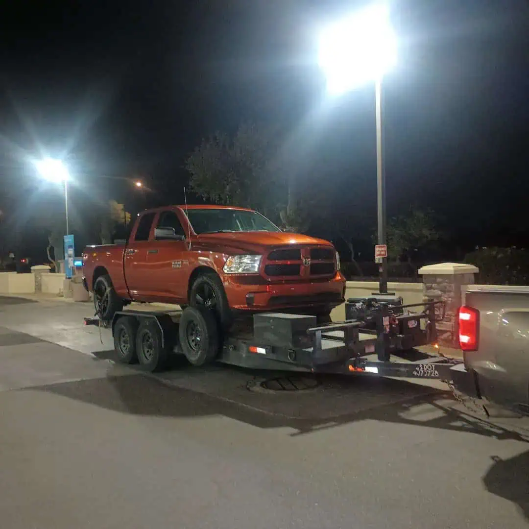 A red pickup truck is loaded onto a flatbed trailer outdoors at night under bright streetlights in AZ. The scene, likely involving roadside assistance Maricopa County, takes place in a parking lot or rest area.