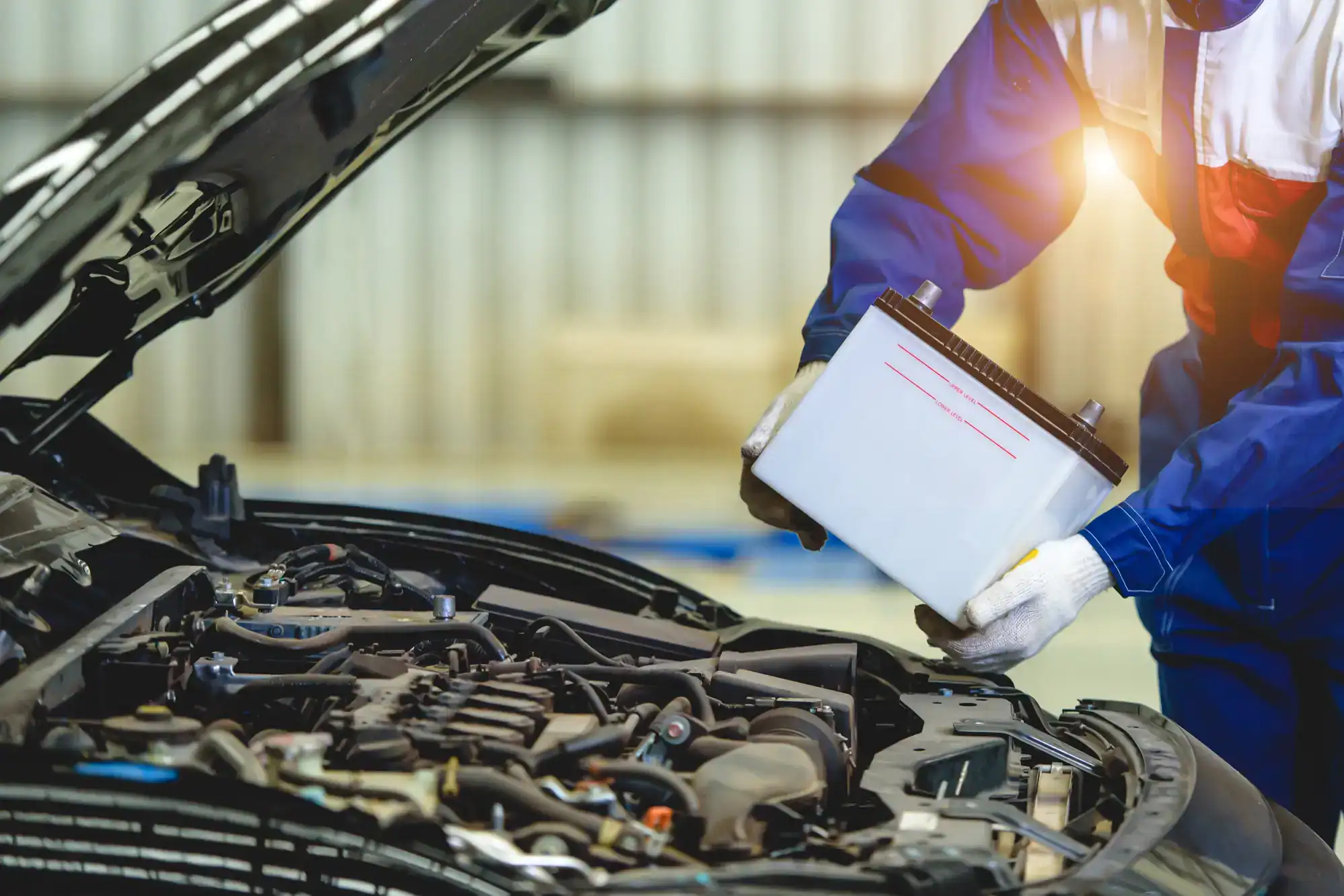 A mechanic in blue coveralls and white gloves is installing or removing a car battery under the hood of a vehicle in a garage, demonstrating the quality care you’d expect from roadside assistance in Maricopa County, AZ. The engine and components are visible.