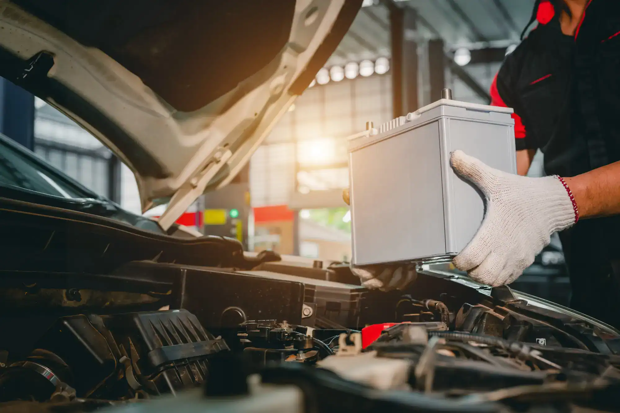 A mechanic wearing white gloves is installing or removing a car battery under the hood of a vehicle in an auto repair shop, offering expert roadside assistance in Maricopa County, AZ.