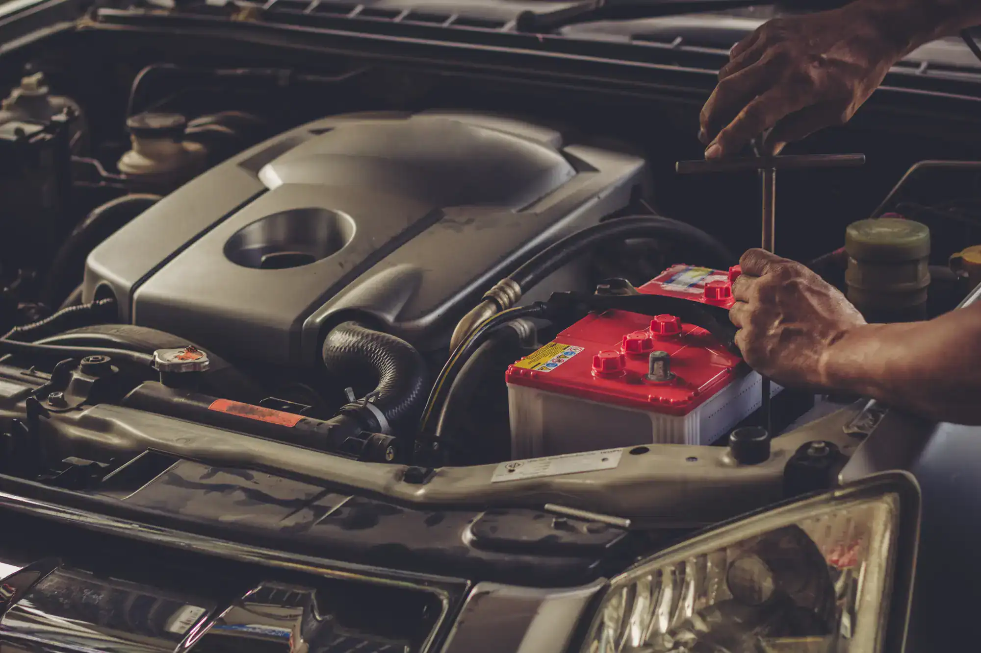 A person’s hands use a tool to work on a car battery under the hood of a vehicle in AZ, surrounded by engine components—ideal for illustrating roadside assistance Maricopa County services.