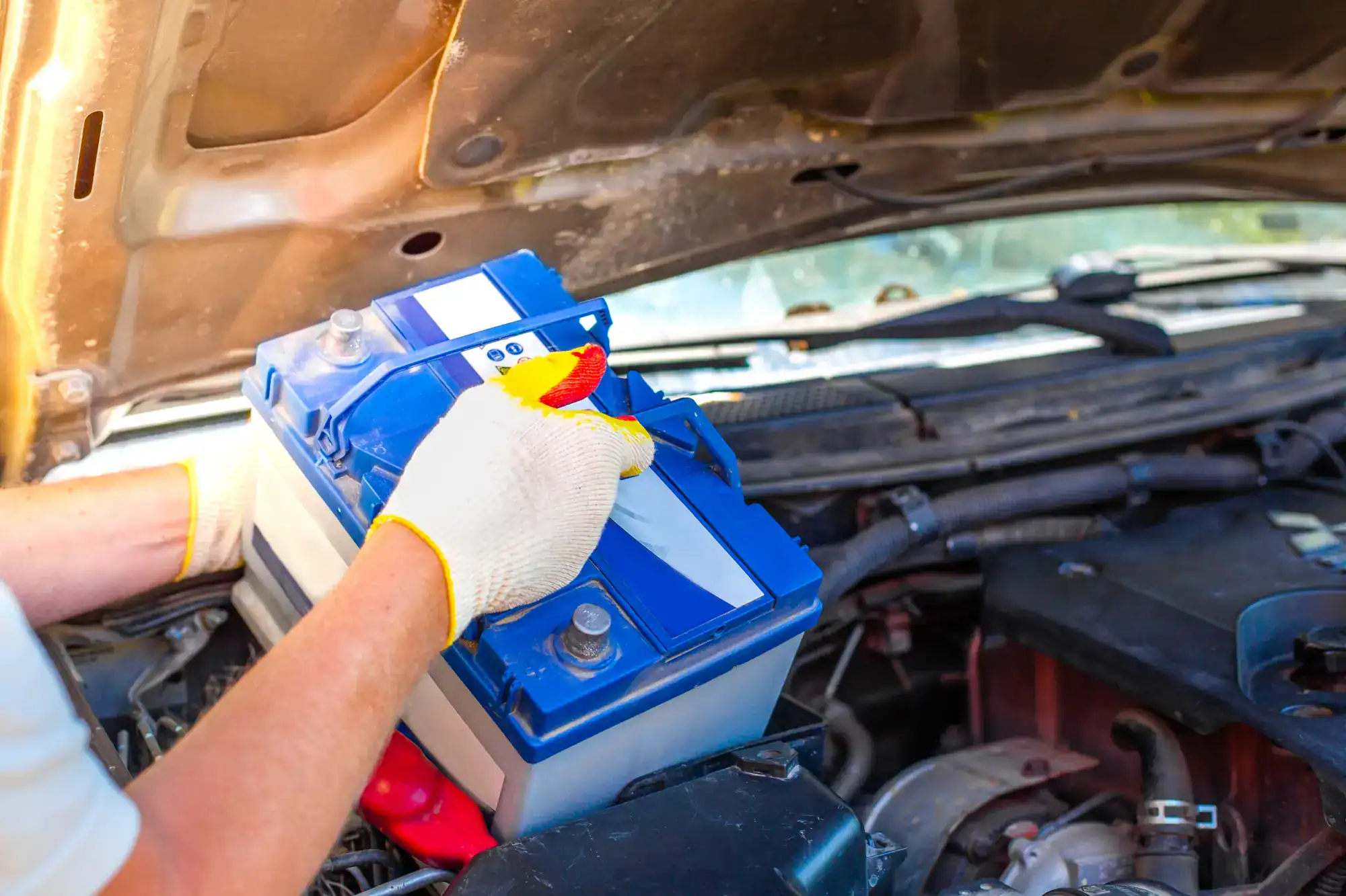A person wearing gloves uses a wrench to install or remove a blue car battery under the raised hood of a vehicle, demonstrating roadside assistance in Maricopa County, AZ.