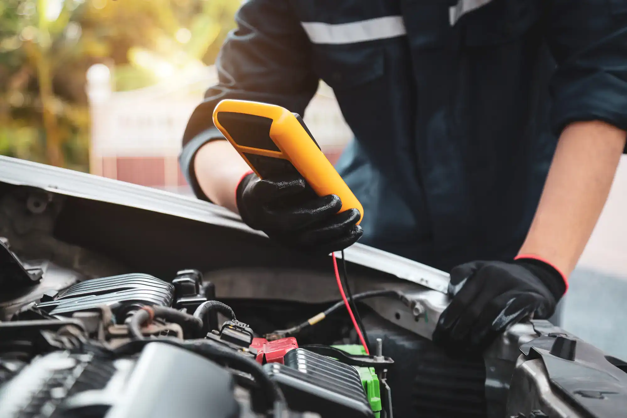 A mechanic wearing black gloves uses a digital multimeter to check a car battery under the open hood of a vehicle, providing expert roadside assistance in Maricopa County, AZ.