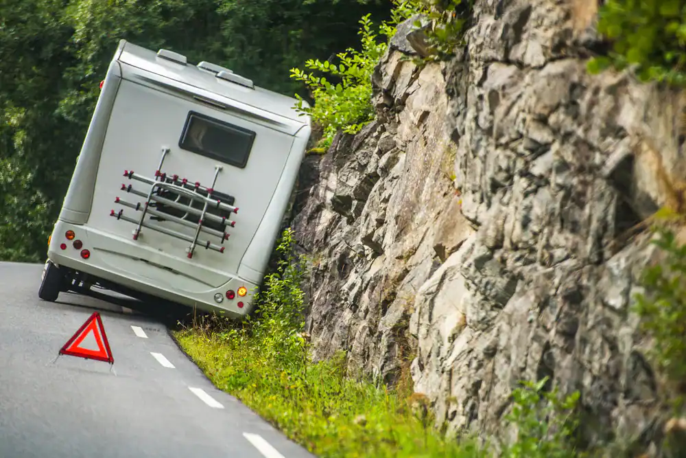 A white camper van is tilted against a rocky cliff on the side of a winding road, with a red warning triangle placed on the road behind it. Trees and greenery surround the area.