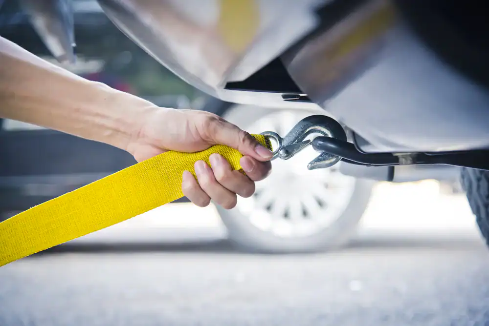 A hand attaches a yellow tow strap with a metal hook to the back of a car, preparing for towing. A blurred car wheel is visible in the background.