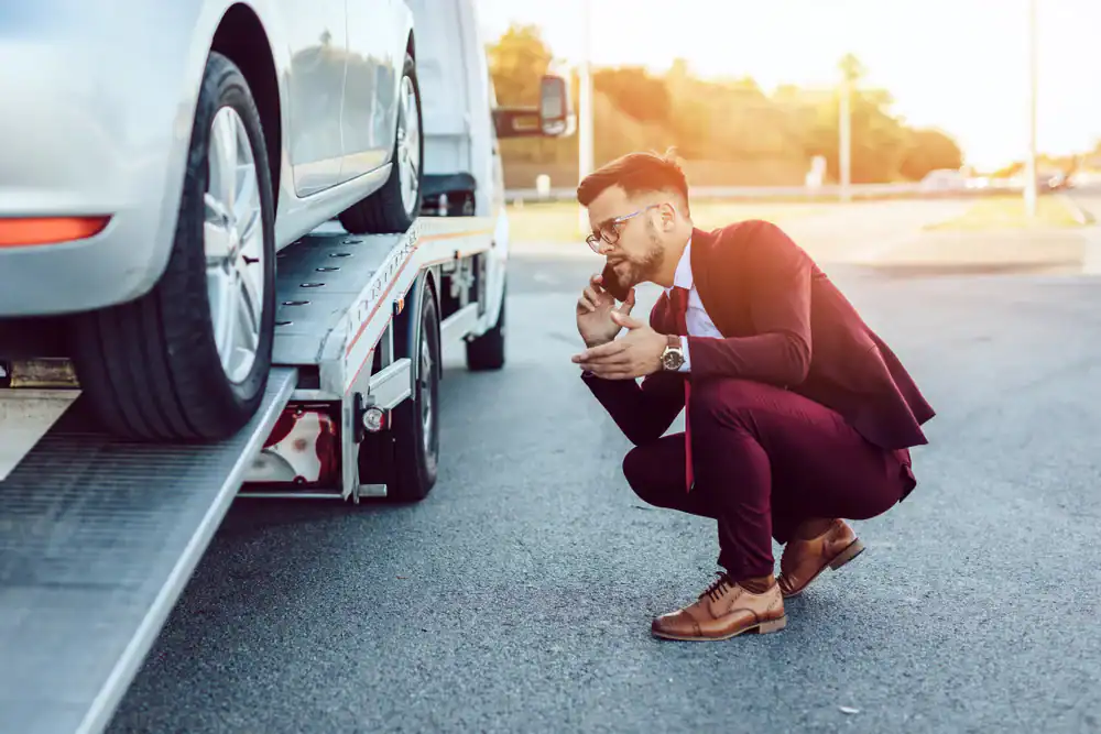 A man in a suit crouches beside a car being loaded onto a tow truck, talking on his phone and looking concerned. The scene is outdoors on a sunny day.