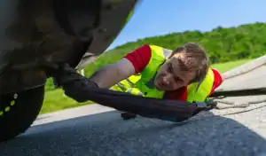 A man in a yellow safety vest kneels on the ground, attaching a tow strap to the underside of a car on a roadside, with green trees and grass in the background.