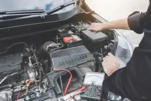 A person checks a car battery under the hood with a multimeter, holding a probe to the battery terminal. Various engine components and wires are visible.