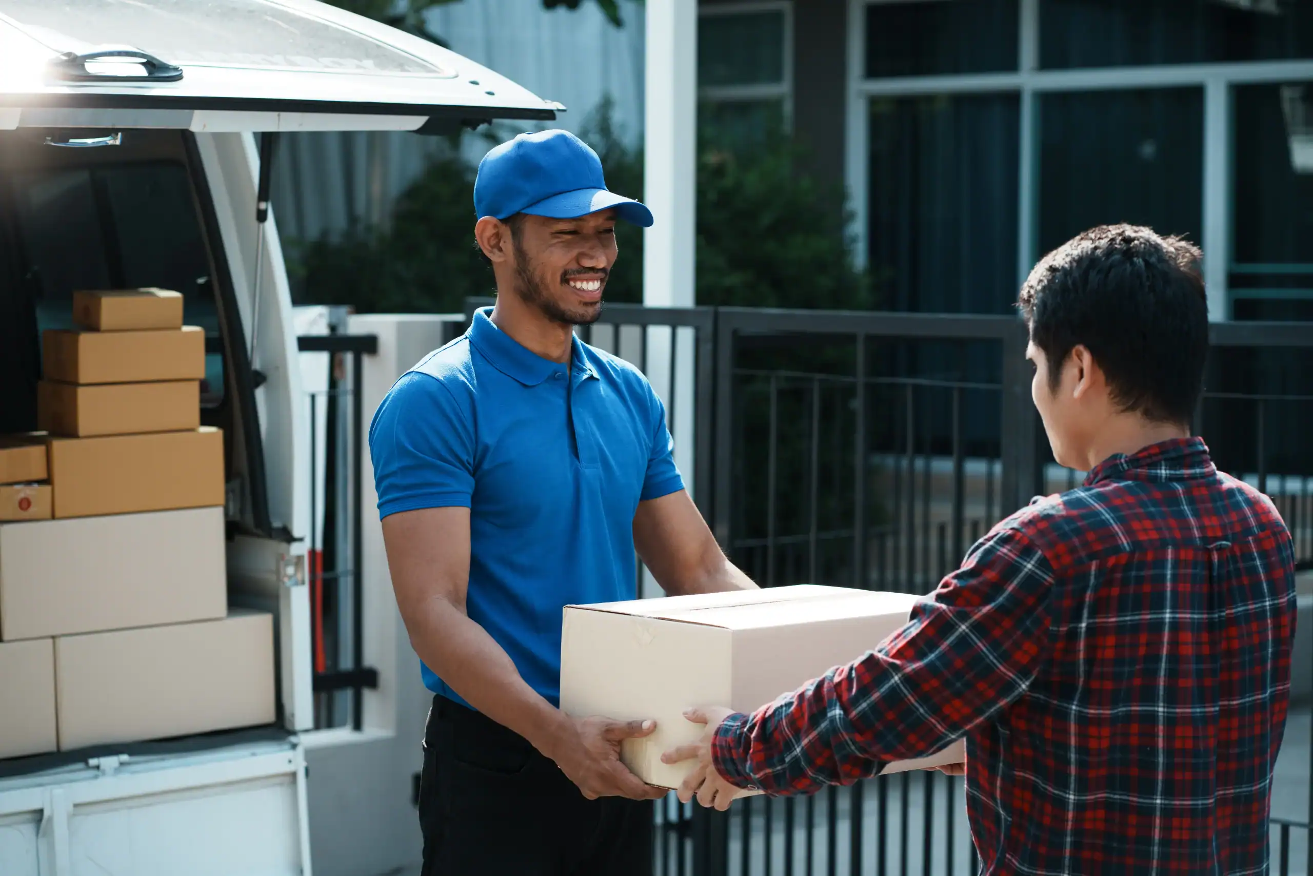 A smiling delivery person in a blue uniform hands a package to a customer in a red plaid shirt outside a building, with a white van full of boxes in the background.