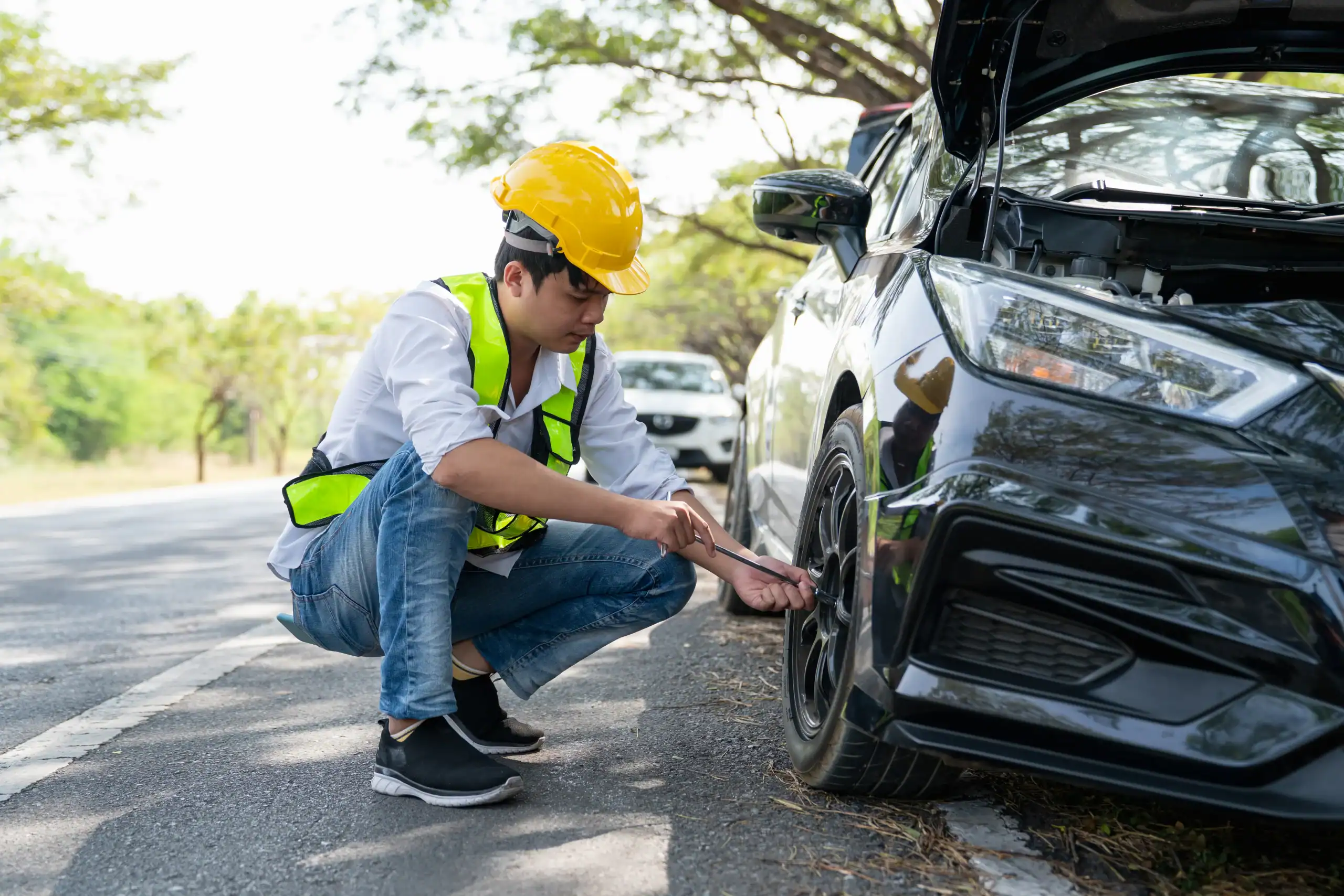 A man wearing a yellow safety helmet, reflective vest, and jeans is crouched by a black car on the roadside, changing a front tire with the hood open. Trees and another car are visible in the background.