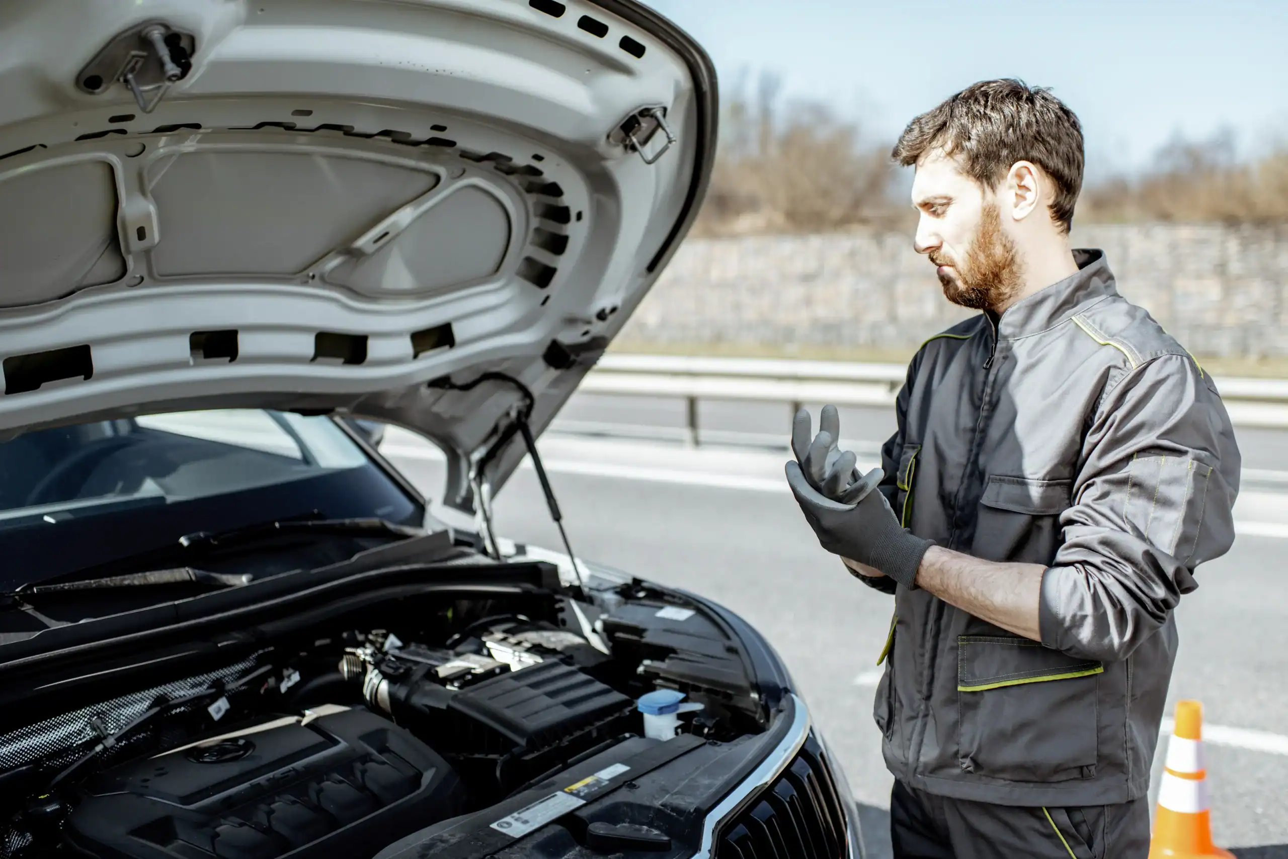 A mechanic in a gray uniform and black gloves stands by a car with its hood open on the side of a road, looking at the engine, with an orange traffic cone nearby.