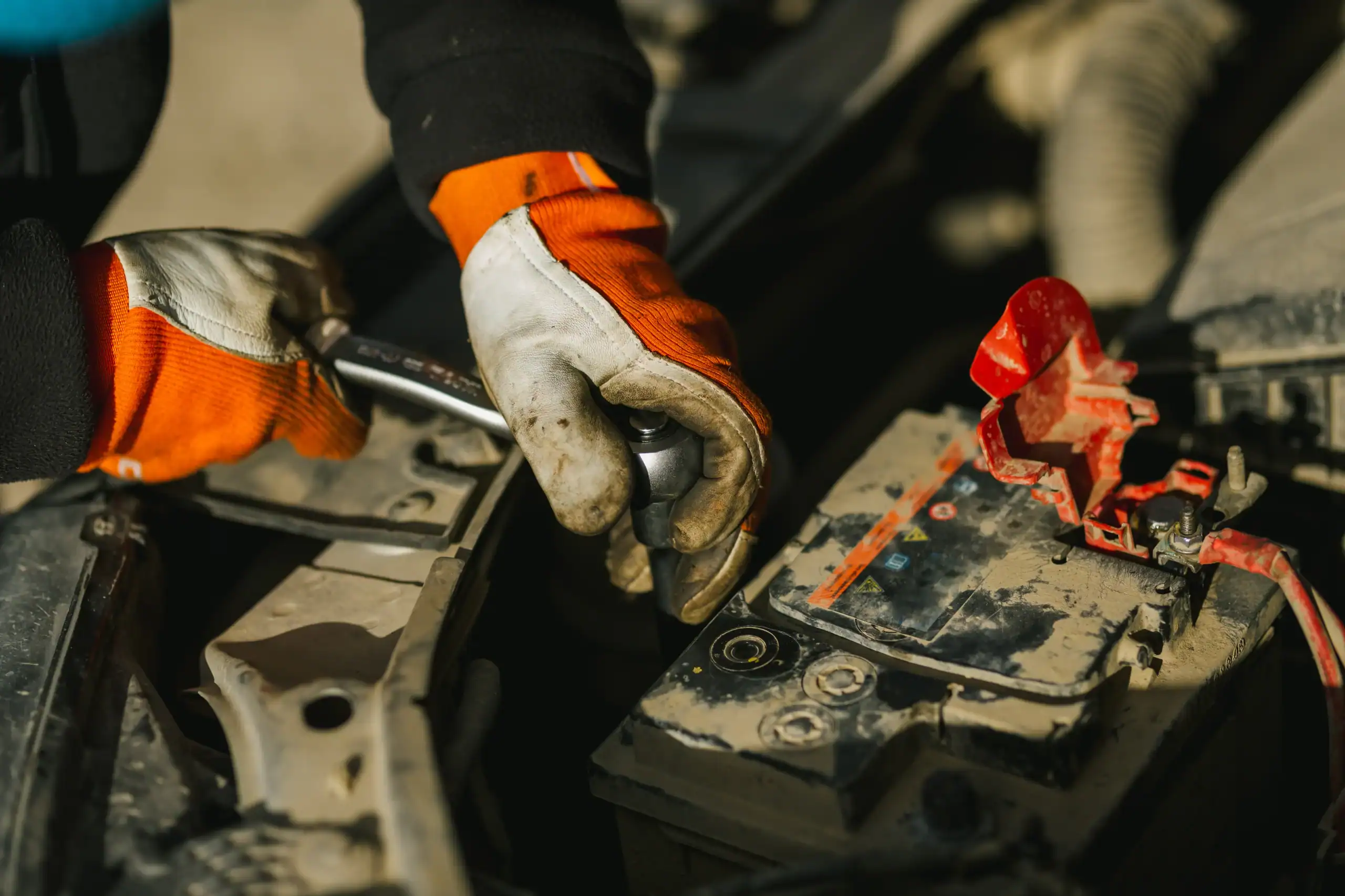 A person wearing orange and white gloves uses a wrench to work on a car battery under the hood; the battery is dusty and has a red terminal cover lifted.