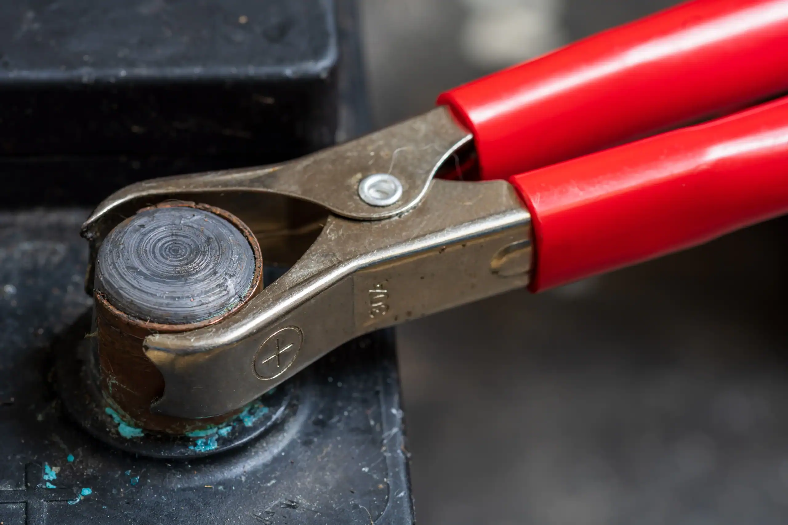 Close-up of a red-handled metal jumper cable clamp attached to the corroded positive terminal of a car battery. The terminal shows signs of oxidation with a blue-green residue.